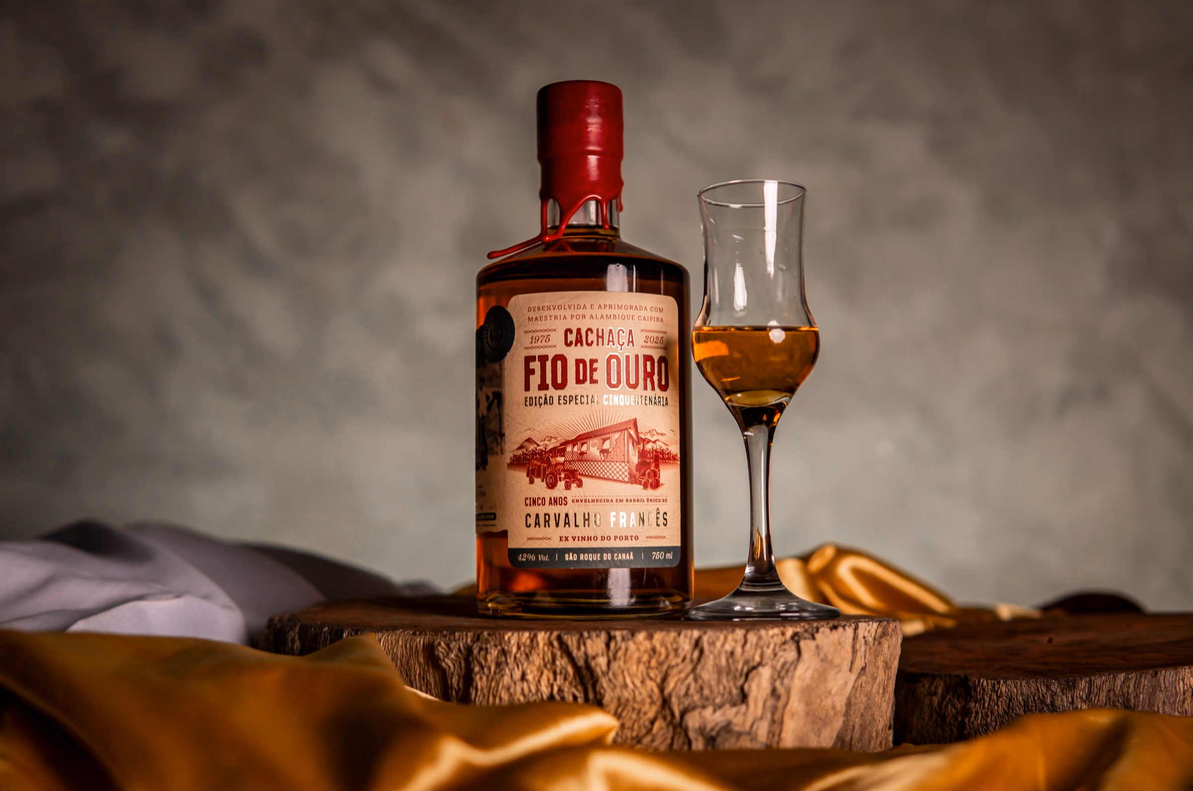 Garrafa da Cachaça Cinquentenária com taça de degustação sobre madeira e cetim dourado